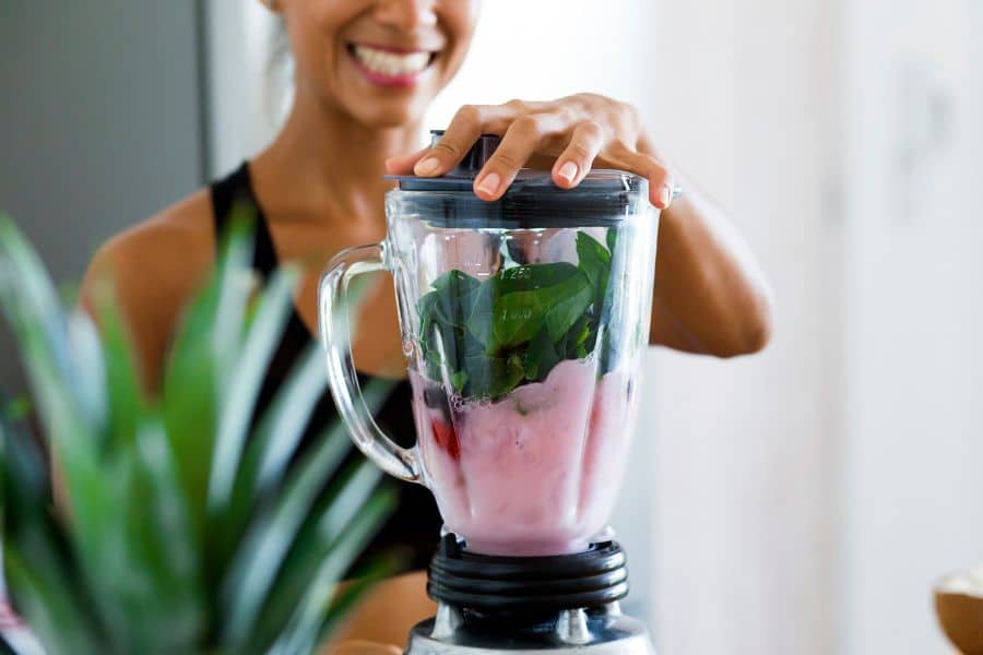 A woman blending a smoothie with leafy greens and pink yogurt in a blender, smiling in the background — highlighting healthy habits often associated with The Weight Loss Trap.