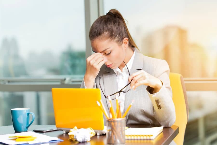 A stressed businesswoman pinching the bridge of her nose while working at her desk, symbolizing emotional triggers that contribute to The Weight Loss Trap.