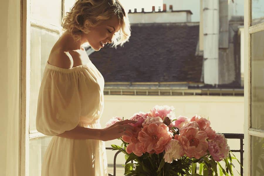 A woman in a soft off-shoulder dress admires fresh pink peonies on a sunny balcony, with Parisian rooftops in the background. A simple pleasure like this helps explain Why French Women Over 50 Look and Feel Younger.