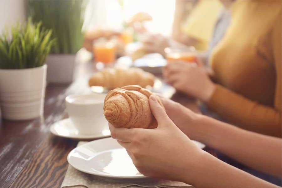 A close-up of a woman’s hands holding a croissant at a café breakfast table, surrounded by coffee, juice, and natural light. Balanced indulgence plays a role in Why French Women Over 50 Look and Feel Younger.