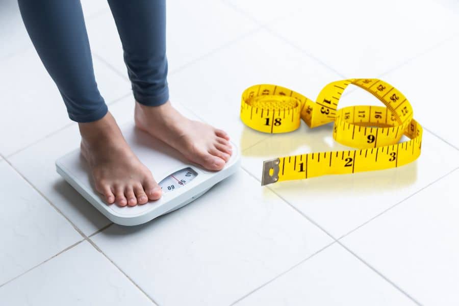 Person standing on a bathroom scale next to a yellow measuring tape, capturing the constant monitoring and pressure behind The Weight Loss Trap.