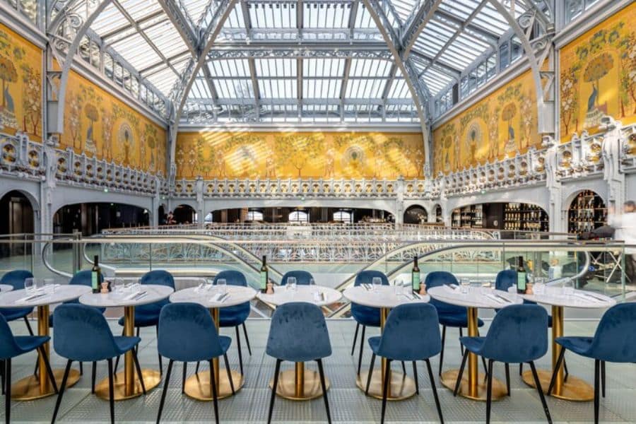 A striking modern café inside a converted historic building showcases bold yellow art nouveau murals, blue velvet chairs, and a glass ceiling. This blend of contemporary and classic makes it one of the most unique Beautiful and Historic Cafés in Europe.