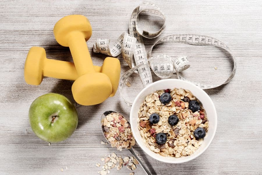 Flat lay of healthy items including a bowl of granola with blueberries, green apple, yellow dumbbells, and measuring tape — representing the well-intended routines that can feed into The Weight Loss Trap.