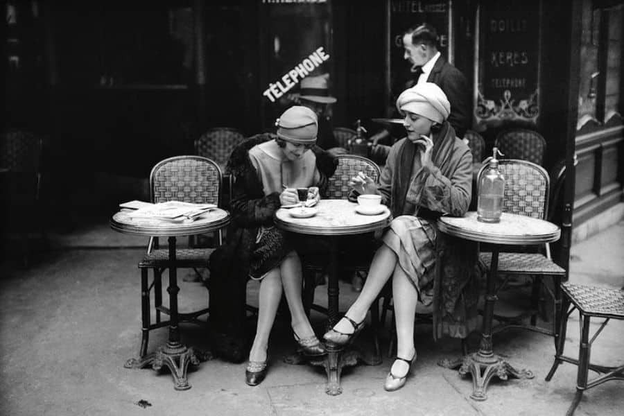 A vintage black-and-white photo shows two fashionable women enjoying coffee at a Parisian café in the early 20th century. Timeless elegance is a central theme in Why French Women Over 50 Look and Feel Younger.