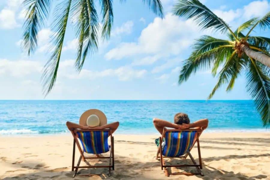 Two people relax on striped beach chairs under palm trees facing the ocean on a sunny day. A peaceful tropical setting that represents the reward of using smart Hacks For Getting Cheaper Vacations.