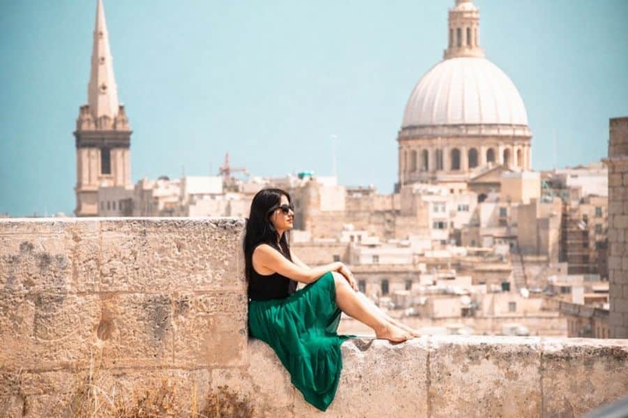 A woman sitting on a stone wall overlooking a domed cathedral and historic skyline, capturing the timeless beauty of European capital cities. The warm stone architecture and clear blue sky highlight the Mediterranean charm of this iconic destination.