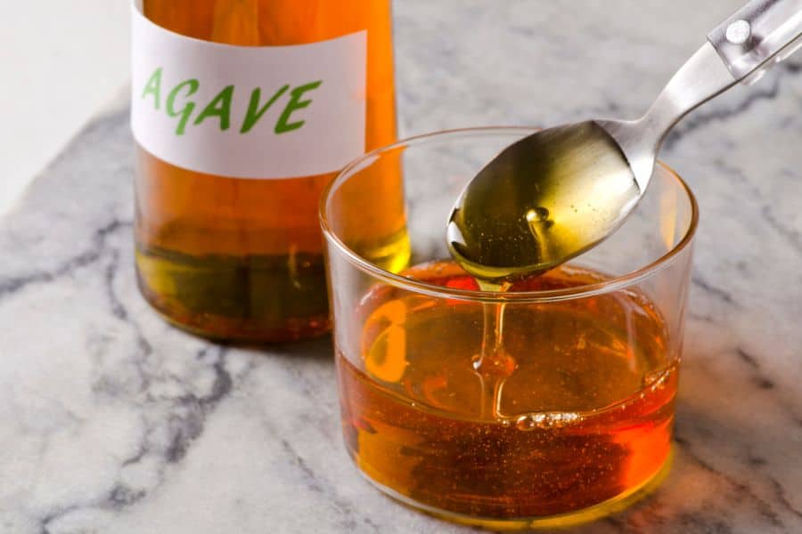 Glass bowl of golden agave syrup with a spoon drizzling syrup and a bottle labeled Agave in the background. This image shows a sweetener often included in Breakfast Staples Triggering Inflammation due to its high fructose content.