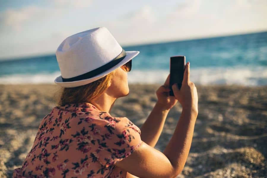 A woman in a white hat and floral shirt takes a photo of the ocean with her phone while sitting on a pebbled beach. Capturing travel memories without overspending is one of the Hacks For Getting Cheaper Vacations.