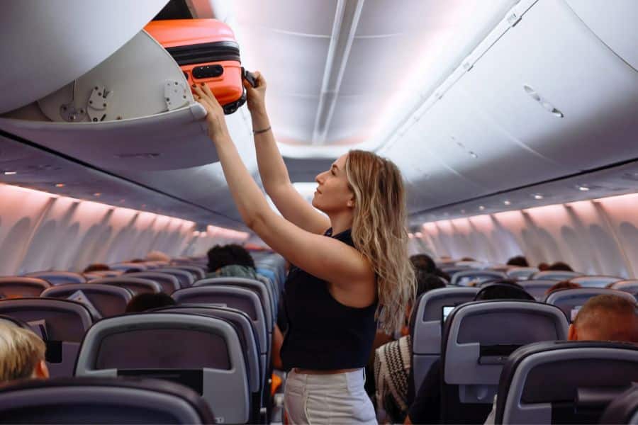 A woman places a bright orange carry-on into the overhead bin of an airplane cabin. Traveling light with a carry-on is one of the best Hacks For Getting Cheaper Vacations to avoid baggage fees.