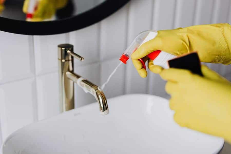Person wearing yellow cleaning gloves spraying a faucet with cleaner in a white bathroom sink. The action highlights how common household cleaning products may contain harmful toxins that affect indoor air and surfaces.