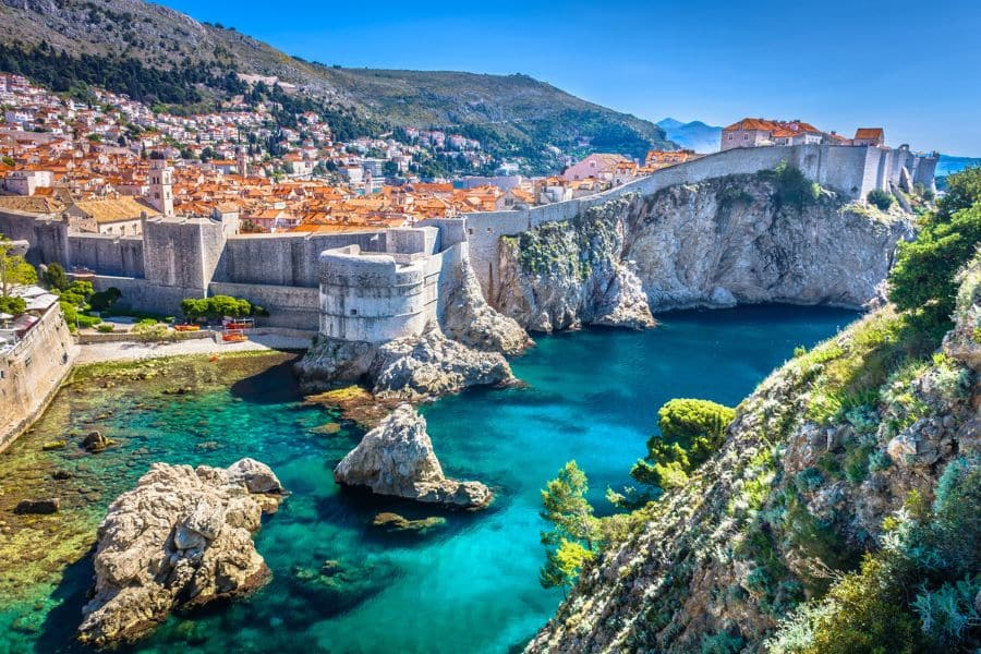 The ancient stone walls of Dubrovnik overlook a rocky coastline and turquoise waters, with red-roofed buildings nestled into the hillside. Dubrovnik is one of the most walkable cities in Europe, ideal for exploring medieval streets and stunning views on foot.