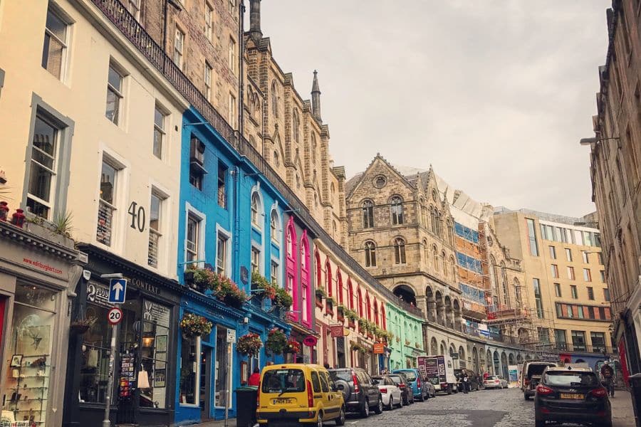 A row of brightly painted shops lines Victoria Street in Edinburgh, curving upward toward grand stone buildings under a cloudy sky. With its compact layout and rich history, Edinburgh is one of the most atmospheric walkable cities in Europe.
