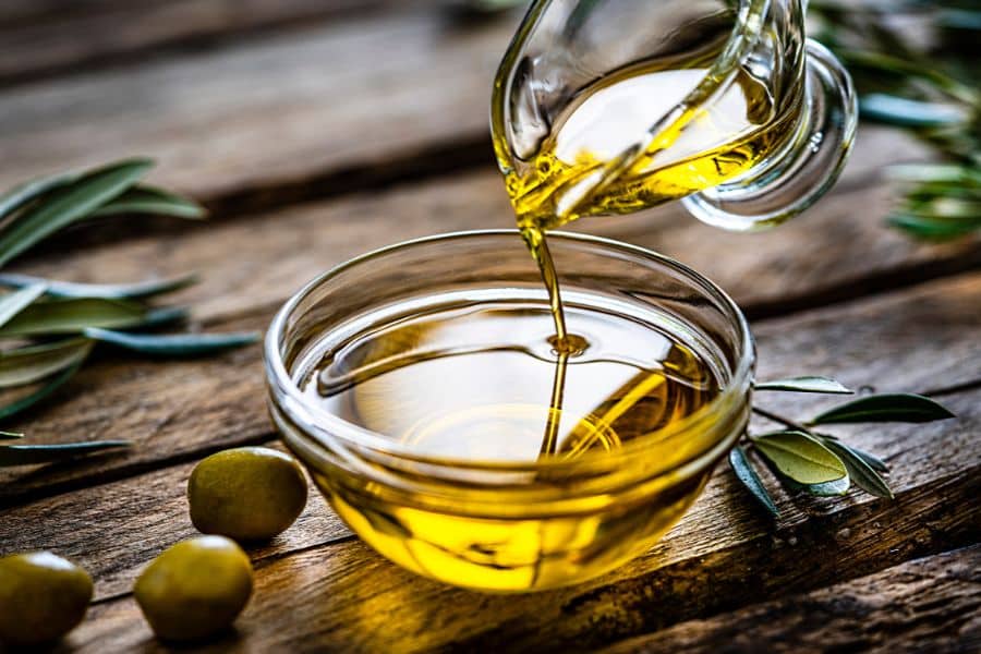 Golden olive oil flows from a glass container into a clear bowl on a wooden table, surrounded by green olives and olive branches. Olive oil is a Mediterranean staple and one of the most effective foods that can reverse aging due to its anti-inflammatory properties.