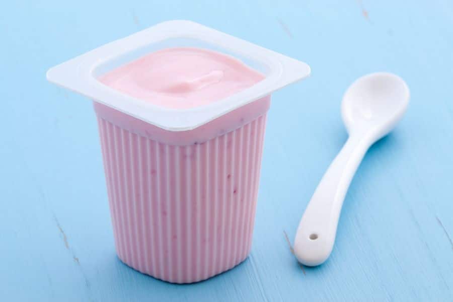 Single serving cup of pink fruit flavored yogurt with a white plastic spoon on a blue surface. Flavored yogurts like this are common Breakfast Staples Triggering Inflammation when they contain added sugars and artificial flavoring.