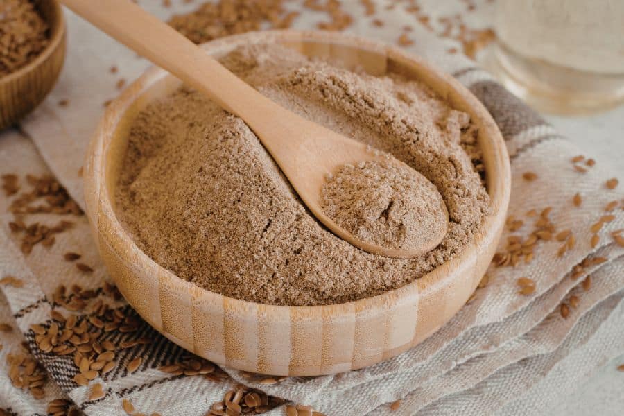 A wooden bowl filled with flaxseed powder and a spoon resting on top, surrounded by scattered flaxseeds. High in fiber and omega-3s, flaxseed is commonly used to support hormone balance and reduce weight gain.