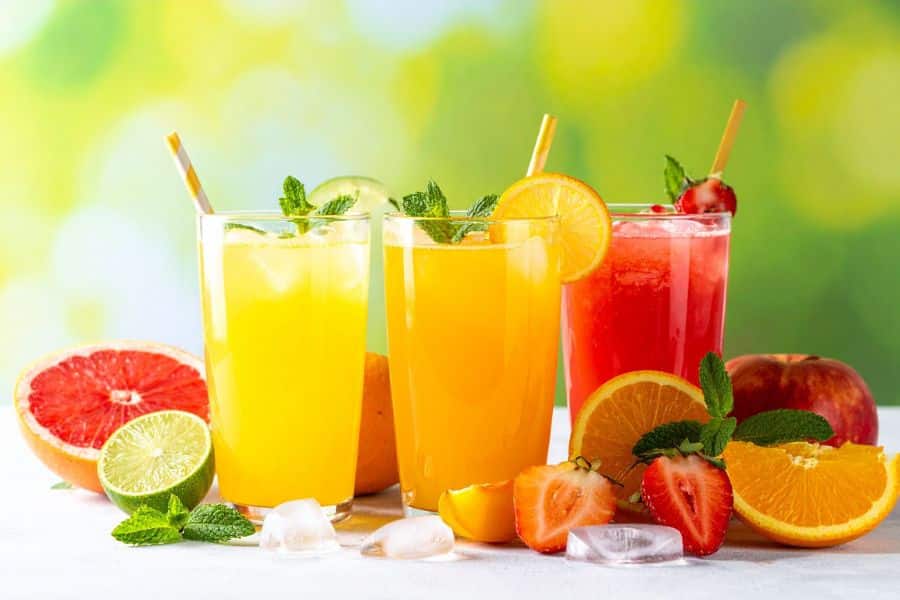Three colorful fruit juices in tall glasses surrounded by sliced citrus, strawberries, and ice cubes. Even fresh looking juices can be Breakfast Staples Triggering Inflammation due to their high natural sugar concentration without fiber.