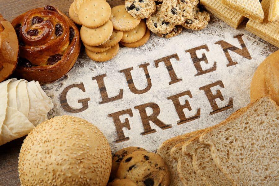 Assortment of bread, cookies, crackers, pastries, and rolls arranged around the words Gluten Free written in flour. Many refined grain products marketed as gluten free can still be Breakfast Staples Triggering Inflammation because they are highly processed.