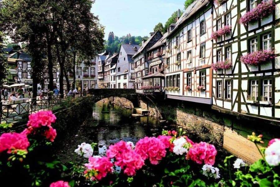 A stone bridge crosses a tranquil stream surrounded by half-timbered houses adorned with pink flowers in Monschau, Germany. This serene view captures the quaint beauty of historic villages in Europe.