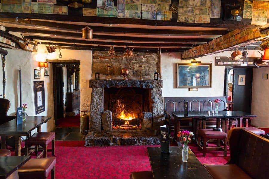 A rustic pub interior with exposed beams, a roaring stone fireplace, and red carpet evokes centuries of hospitality tradition. This inviting setting reflects the old-world charm typical of historic villages in Europe.