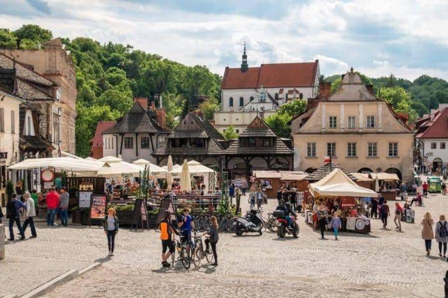 A lively market square bustles with people, outdoor stalls, and bicycles against a backdrop of historic buildings and a hilltop church in Kazimierz Dolny, Poland. The vibrant scene is a perfect example of life in historic villages in Europe.
