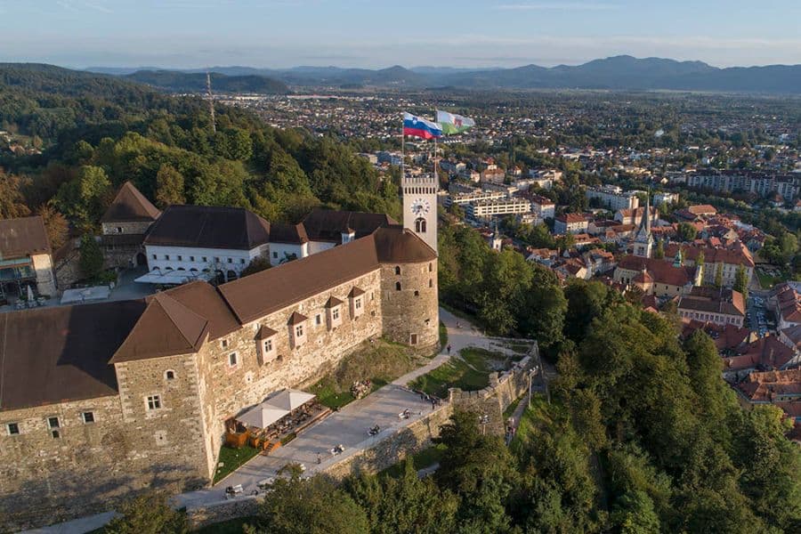 An aerial view of a hilltop castle flying a national flag above red roofed buildings, representing the historic character of European capital cities. The fortress overlooks a sprawling urban landscape surrounded by green hills.