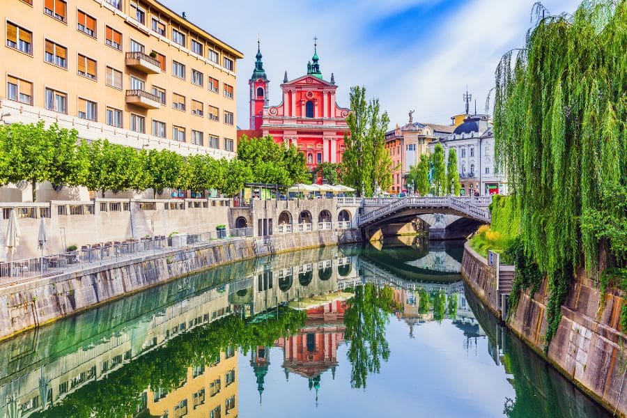 A picturesque canal lined with pastel buildings and a pink church reflected in calm water, showcasing the elegance of European capital cities. A stone bridge arches over the river, adding to the romantic cityscape.