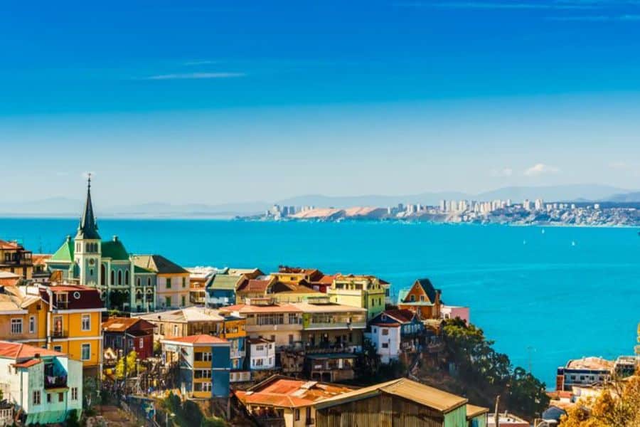 Colorful hillside buildings and a green church overlook a bright blue bay with a distant skyline and mountains. Exploring off-the-beaten-path coastal cities is one of the hidden Hacks For Getting Cheaper Vacations.