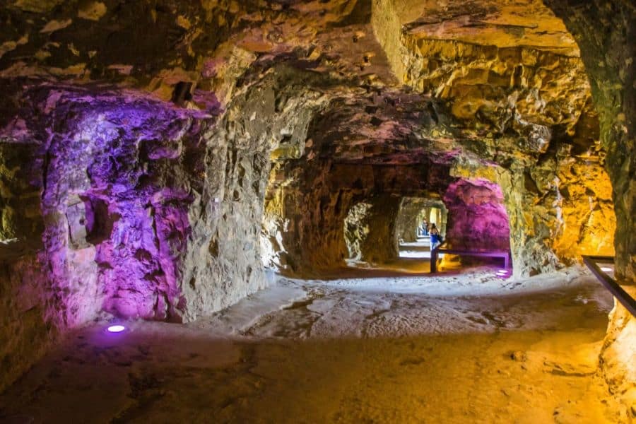 Expansive rock tunnel illuminated with purple and golden lights, highlighting the natural stone formations inside Underground Cities in Europe. The uneven walls and carved pathways show how the space blends geology with human excavation.