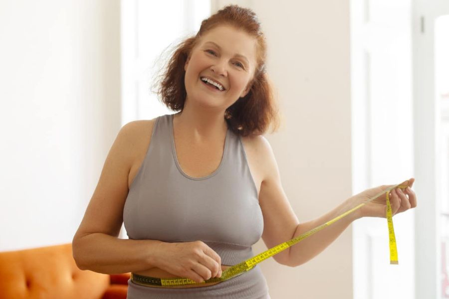 A smiling woman measures her waist with a tape measure in a cozy living room. This visual highlights progress tracking as part of Weight Loss Habits For Women Over 55 that encourage sustainable lifestyle changes.