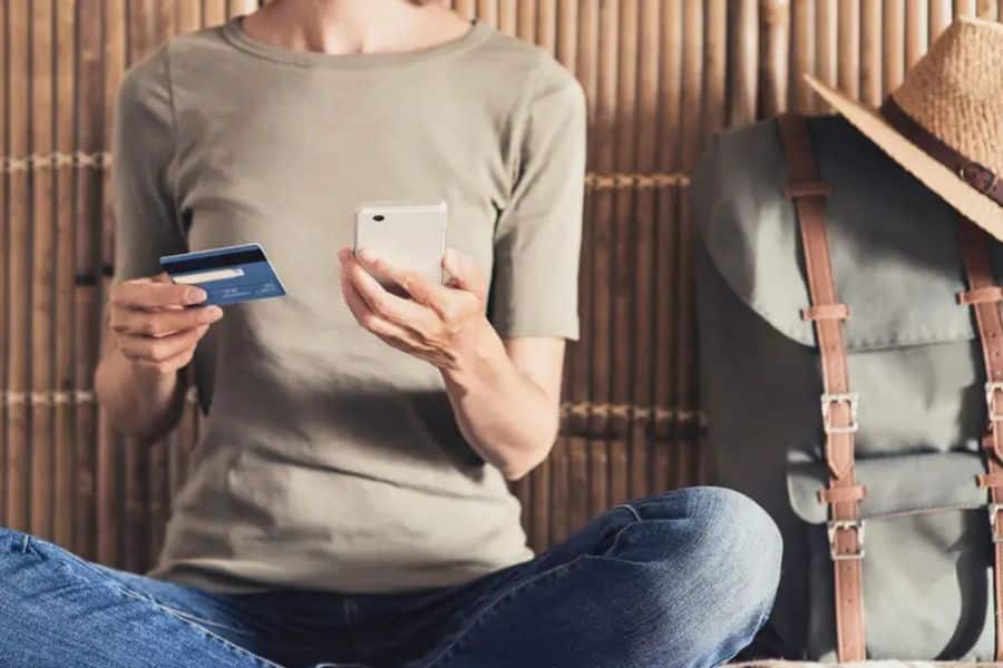 Person sitting cross-legged with a credit card in one hand and smartphone in the other, next to a backpack and sun hat. Booking online with rewards points is one of the digital Hacks For Getting Cheaper Vacations.