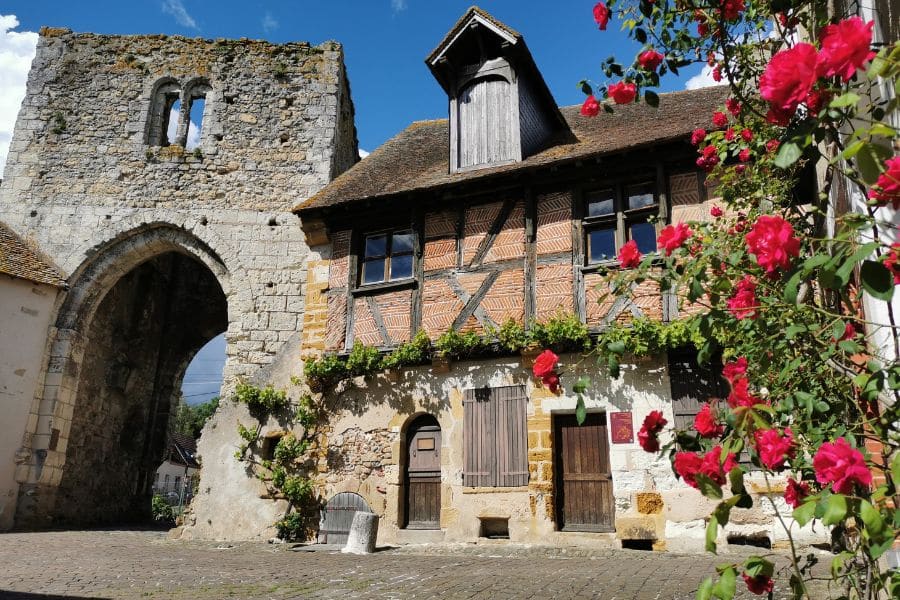 A stone tower and half-timbered house with rustic shutters and rose-covered walls form a quiet courtyard in a French village. The preserved textures and blooms capture the essence of historic villages in Europe.