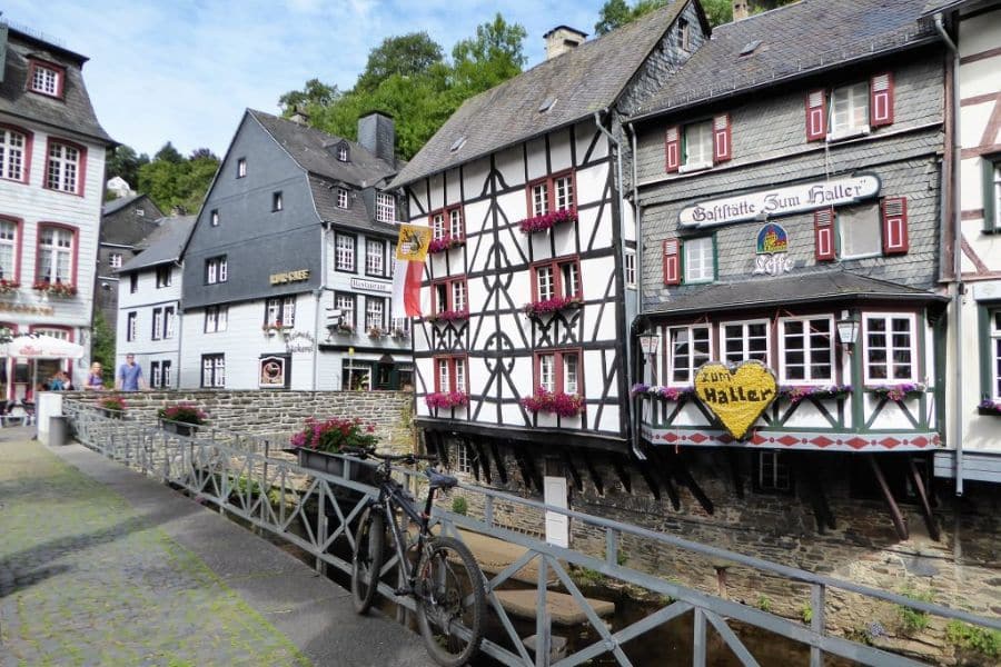 Timber-framed inns with flower boxes line a quaint bridge over a narrow stream in Monschau, Germany. The charming facades and relaxed setting illustrate the timeless appeal of historic villages in Europe.