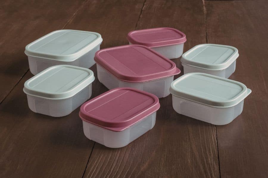Stack of reusable plastic food storage containers with pink and gray lids on a wooden table. The image connects everyday food storage items to potential exposure to harmful toxins from certain plastics.