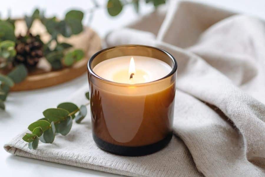 Lit candle in a brown glass jar placed on a soft cloth with greenery in the background. The cozy scene contrasts with concerns that some scented candles may release harmful toxins into indoor air.