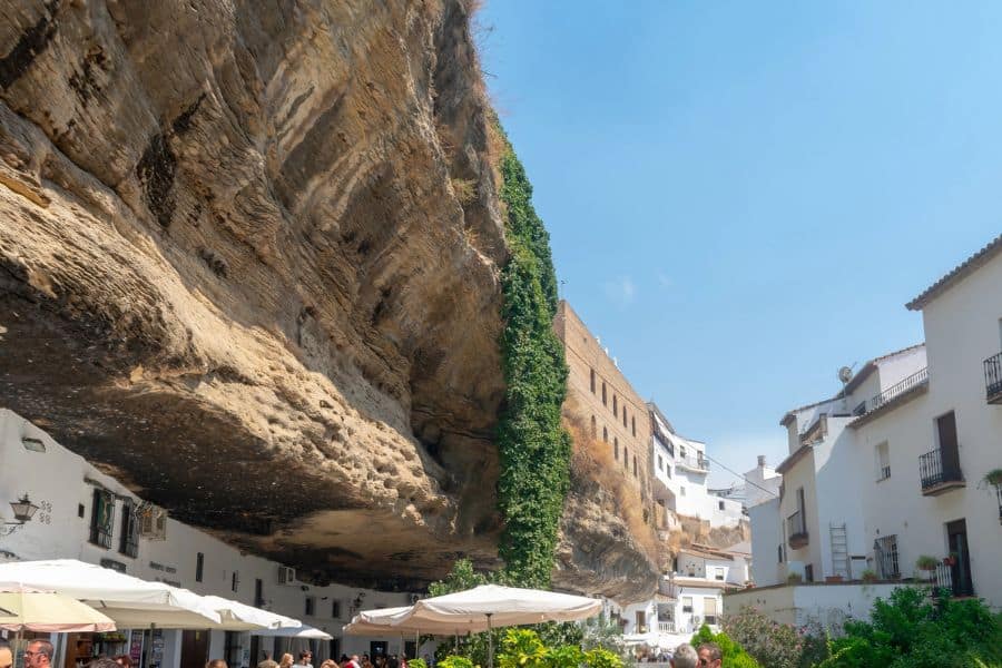 White village houses built beneath a massive rock overhang with outdoor cafes and greenery, demonstrating how surface towns connect to Underground Cities in Europe. The dramatic cliff formation towers above the lively street below.