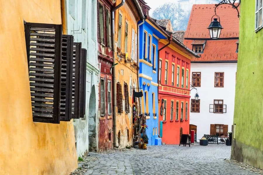 A cobblestone street winds through a row of brightly colored buildings in Sighișoara, Romania, with hues of yellow, blue, red, and green adding vibrance to the medieval village. This lively alleyway captures the character and charm of historic villages in Europe.