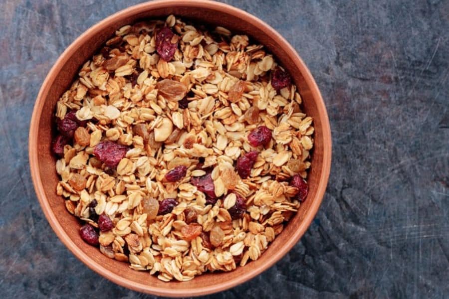 Wooden bowl filled with granola containing oats, raisins, and dried cranberries on a dark surface. Granola is often considered healthy but can be one of the Breakfast Staples Triggering Inflammation when it is high in sugar and refined oils.