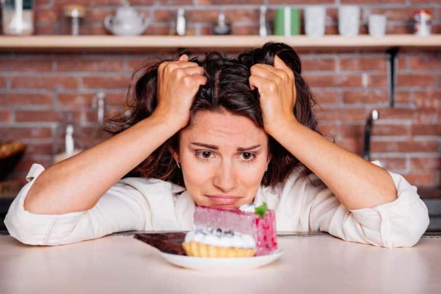 A woman looking stressed while staring at a slice of cake on a table, symbolizing the search for healthier sweetener alternatives. Her expression reflects the challenge of reducing sugar while satisfying cravings.
