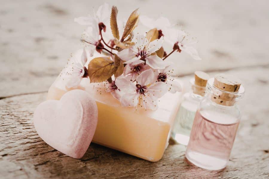 Natural bar of soap topped with delicate pink blossoms beside small glass bottles of bath oil on a rustic wooden surface. The soft floral arrangement highlights gentle, plant based skincare as a soothing alternative to products that may contain harmful toxins.