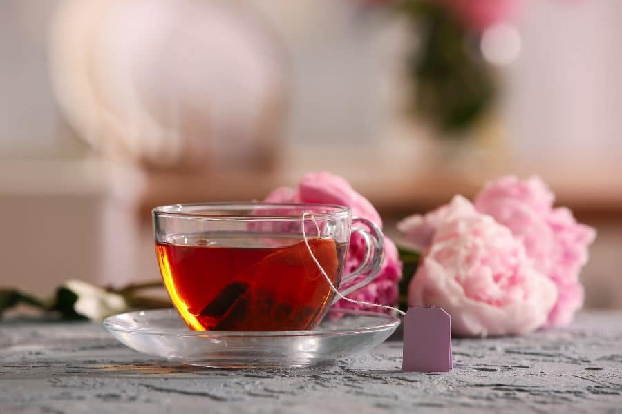 Clear glass cup of red herbal tea with a tea bag resting on a saucer, pink peonies blurred in the background. The calming scene suggests choosing natural beverages to reduce exposure to harmful toxins found in some processed drinks.