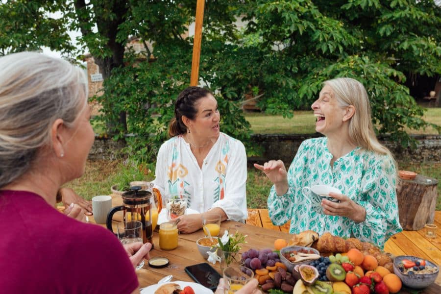 Three women enjoying breakfast outdoors at a wooden table filled with fresh fruit, juice, coffee, and pastries, laughing and talking under the trees. A joyful scene of friends eating breakfast together in a relaxed garden setting.
