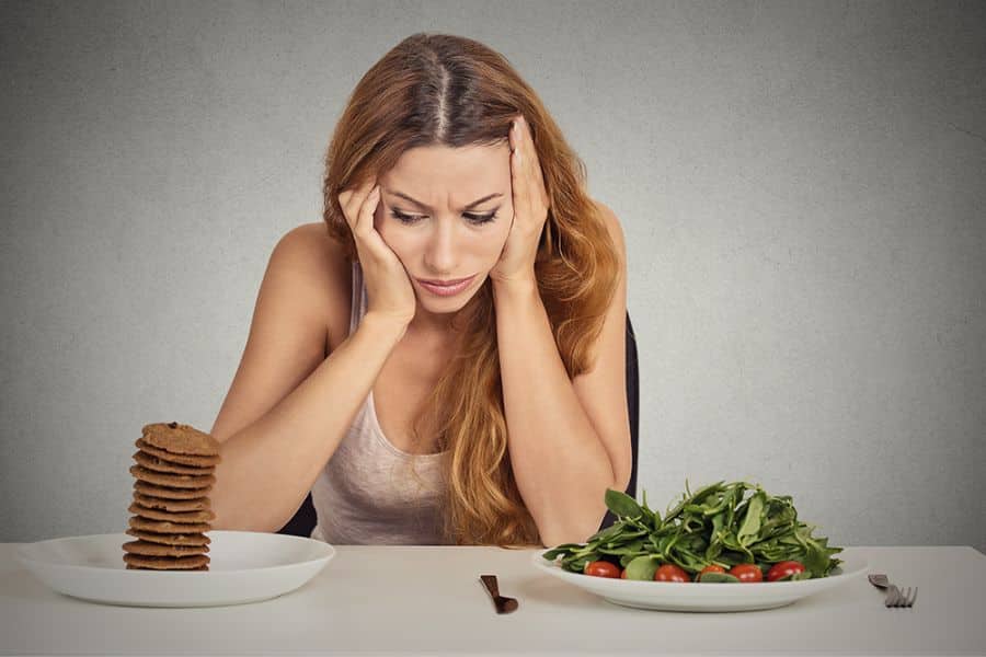 Woman sitting at a table looking stressed while choosing between a plate of cookies and a bowl of fresh salad, reflecting common diet struggles during Weight Loss In Midlife. Her frustrated expression highlights the challenge of balancing cravings and healthy choices.