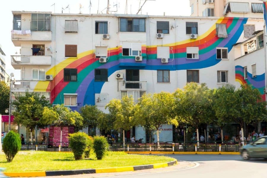 Most Colorful Places in Europe featuring a city apartment building adorned with a large rainbow mural sweeping across its white exterior. The bright stripes of red, orange, yellow, green, blue, and purple transform the urban facade into a vibrant public artwork.