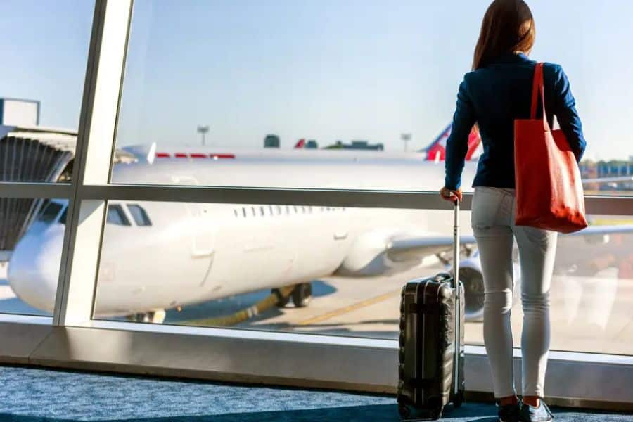 Woman standing at an airport window with a suitcase, gazing at a plane parked at the gate. Flying mid-week or during off-peak hours is one of the top Hacks For Getting Cheaper Vacations.