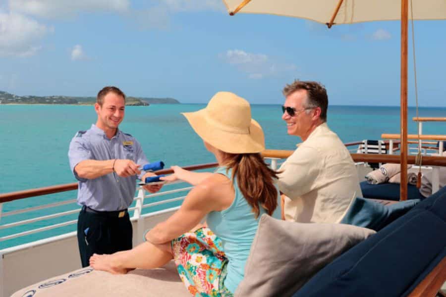 A couple receives towels from a cruise staff member while lounging on a sunny ship deck. Booking last-minute cruise deals is one of the relaxing Hacks For Getting Cheaper Vacations.