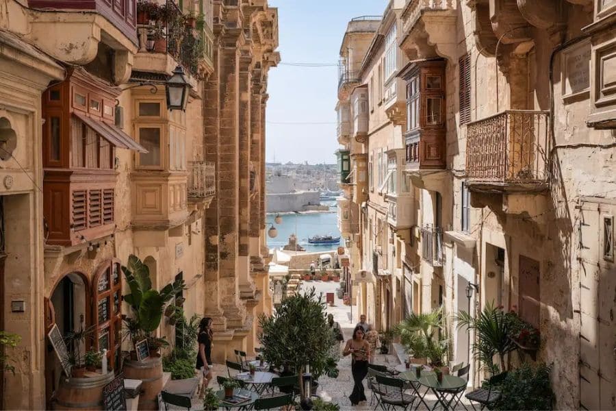 A narrow historic street opening toward a bright harbor view, reflecting the layered architecture common in European capital cities. Balconies, cafes, and stone facades create an inviting urban atmosphere.