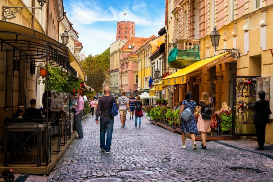 A lively cobblestone street filled with pedestrians, colorful facades, and outdoor cafes, capturing the everyday vibrancy of European capital cities. A historic tower rises in the background above the pastel buildings.