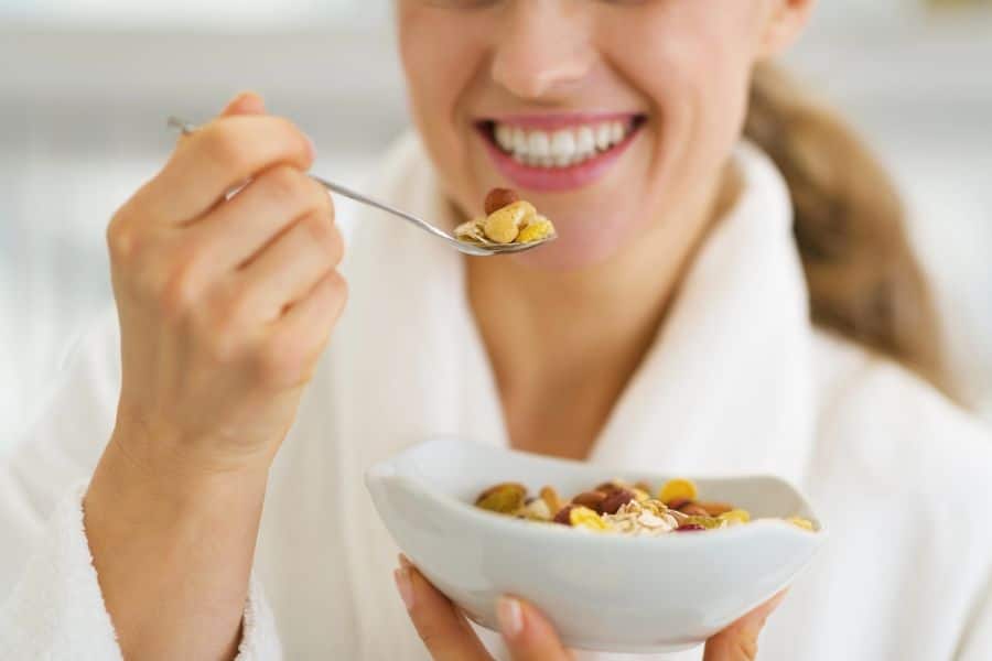 Close-up of a woman smiling as she lifts a spoonful of granola with dried fruit and nuts from a bowl, wearing a white robe. She is mid-bite, clearly enjoying eating breakfast with a healthy cereal mix.