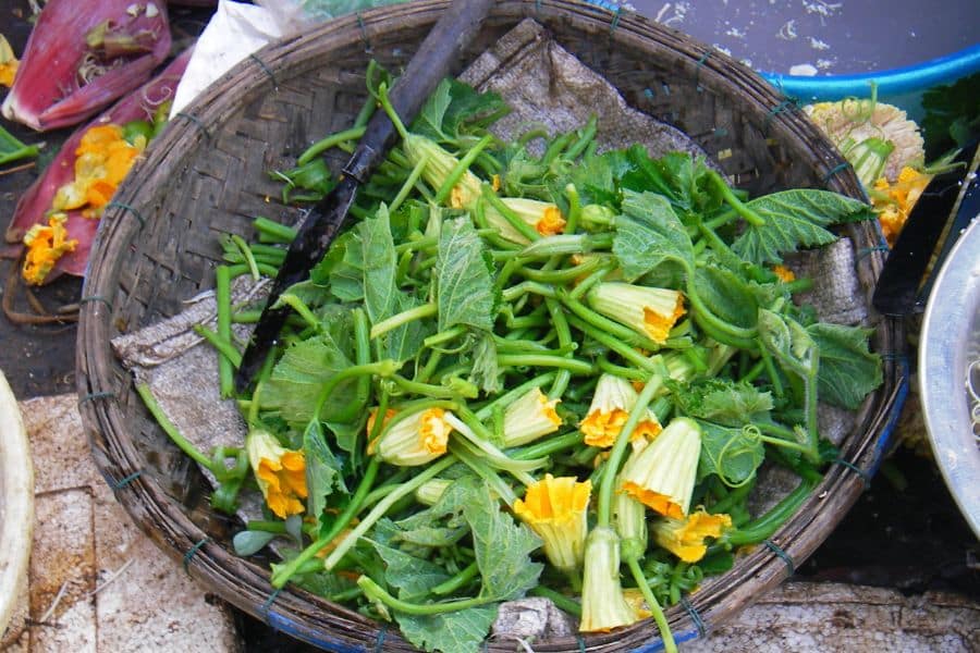 Basket filled with fresh squash shoots, tender stems, and yellow blossoms at an outdoor market. This works well for content about edible leaves because it shows leafy vine tips and flowers that are harvested for cooking.