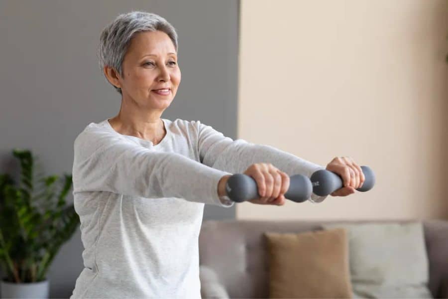 Older woman doing a hand grip test related strength workout at home while holding two gray dumbbells straight out in front of her. She wears a light gray long sleeve shirt and smiles in a living room with a sofa and plant in the background.